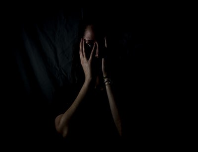 Photo of woman with her hands covering her face in a dark room depicting a fear of something or someone.